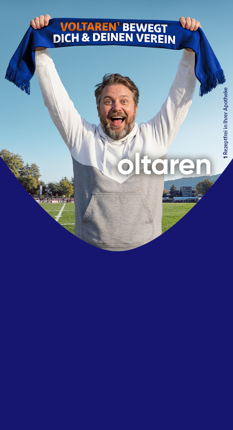 Person steht auf einem Fußballfeld und hält einen blauen Schal mit der Aufschrift 'Voltaren bewegt dich & deinen Verein' über den Kopf; im Hintergrund Spielfeld, Bäume und Zuschauer